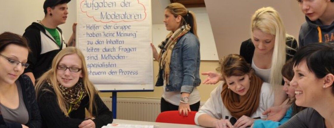 SchülerInnen sitzen um einen Tisch, gestalten ein Plakat, zwei weitere stehen im Hintergrund an einem Flipchart